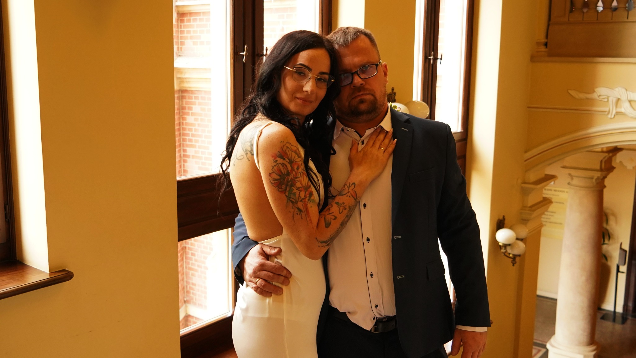 Para w objęciach pozuje na tle klasycznego wnętrza z kolumnami i oknami; kobieta w białej sukience z odkrytymi plecami i tatuażami, mężczyzna w granatowym garniturze.