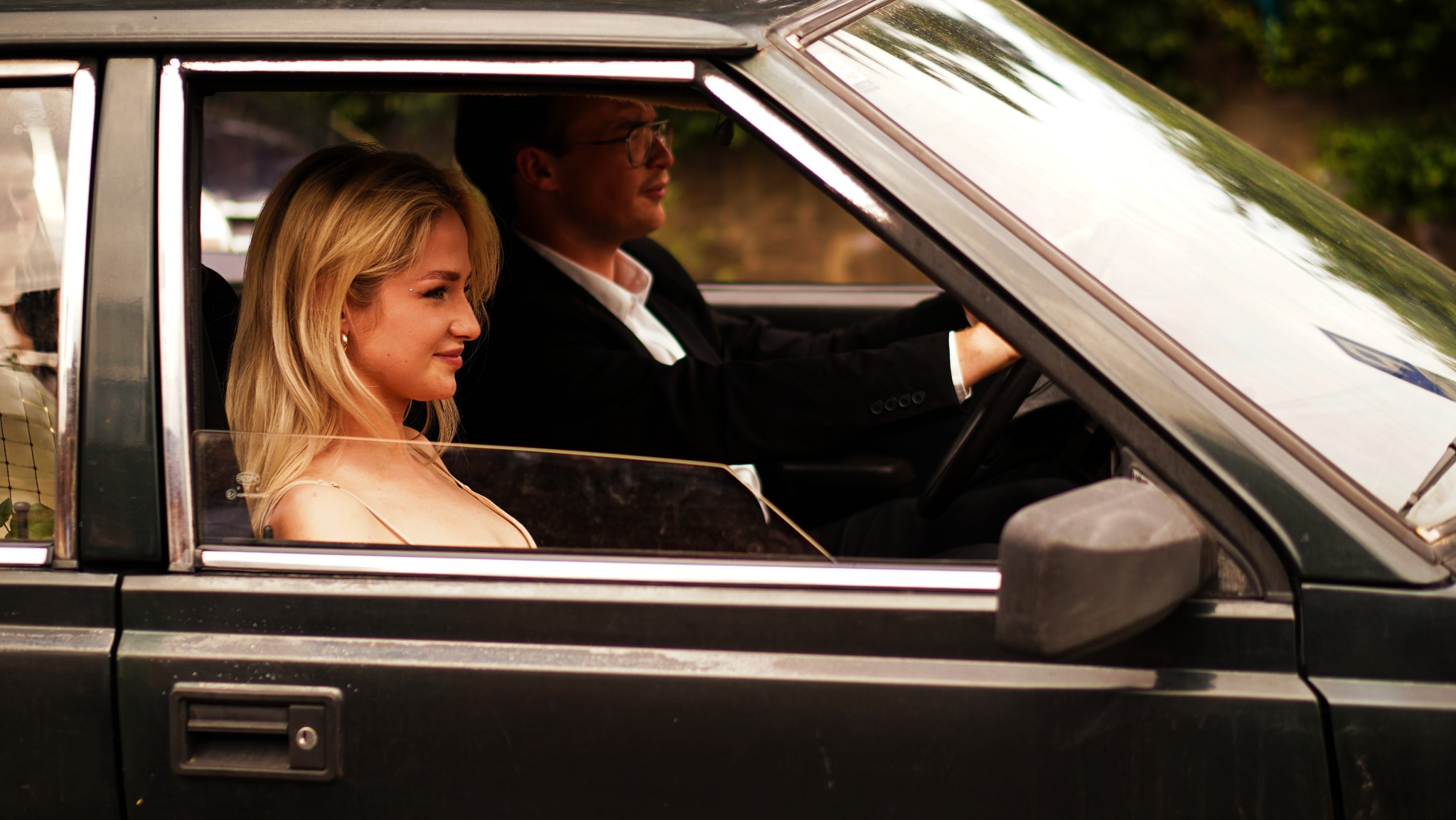 Kobieta z platynowymi blond włosami i subtelnym makijażem w samochodzie, obok mężczyzna w garniturze za kierownicą, ujęcie z zewnątrz przez szybę samochodu.