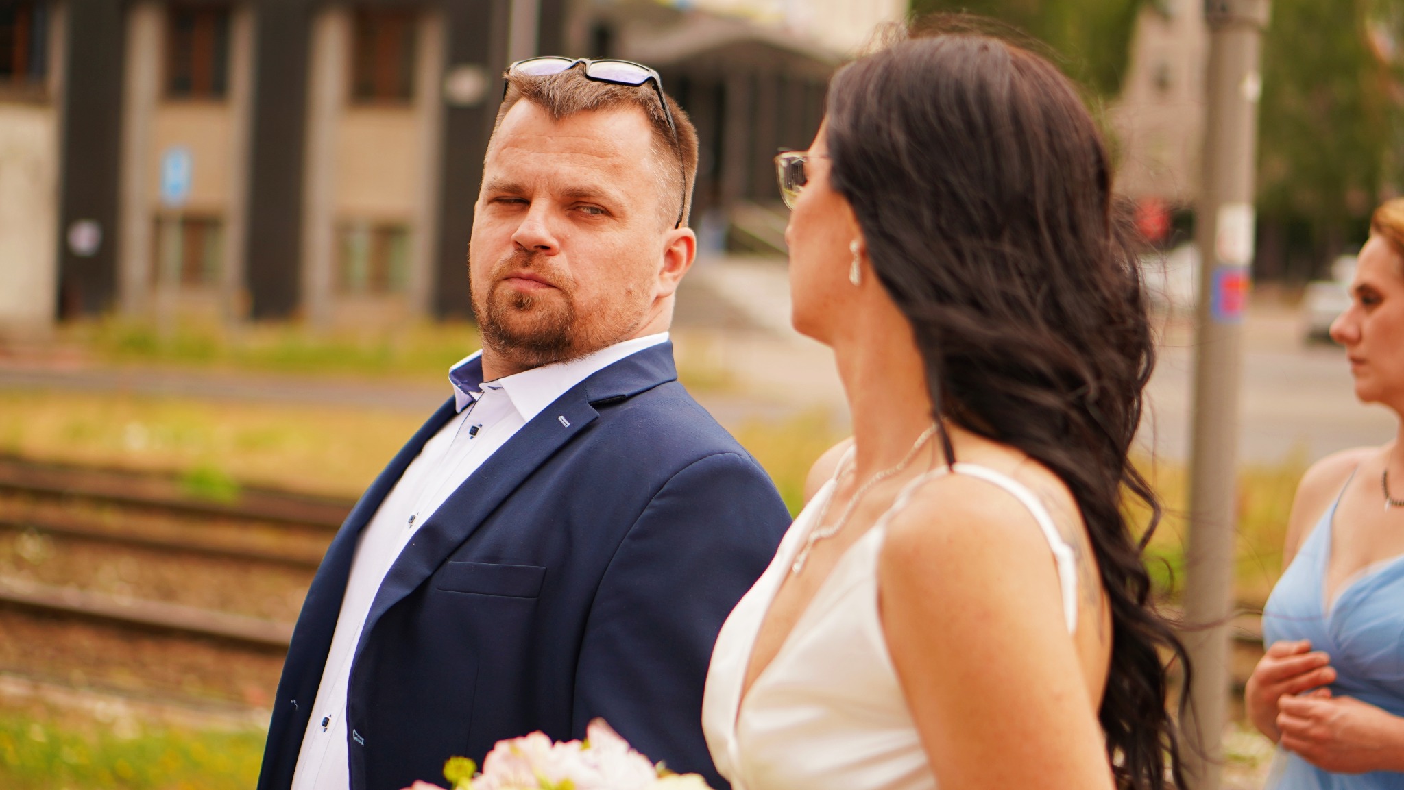 Ujęcie pary młodej w plenerze: mężczyzna w granatowym garniturze z okularami na głowie spogląda w obiektyw, obok kobieta w białej sukni z bukietem, w tle tory kolejowe i budynek.