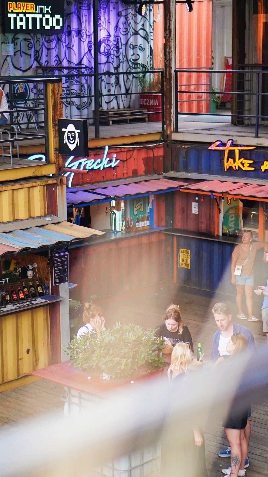 Widok z góry na plac z barami i restauracjami zbudowanymi z kontenerów, z neonowymi szyldami 'Grecki' i 'Take a...' oraz graffiti na ścianach kontenerów. W centrum stolik z grupą osób.