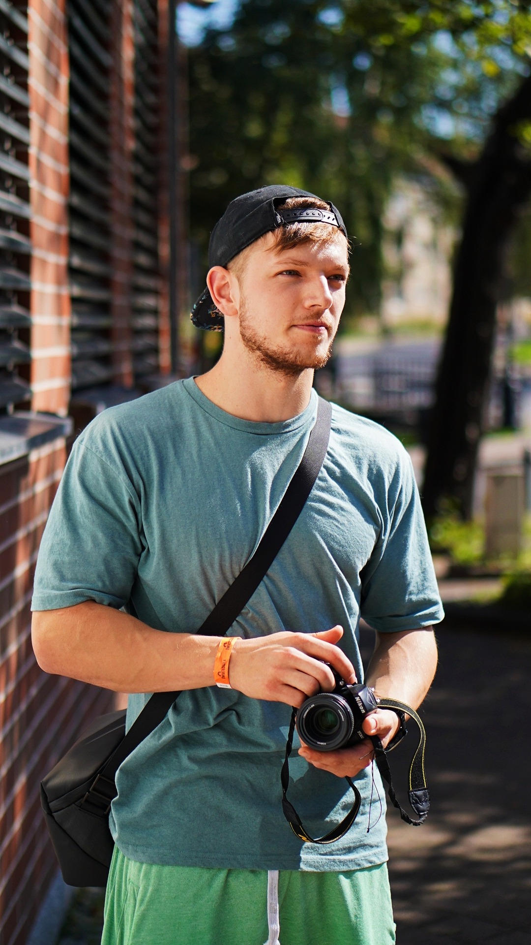 Młody mężczyzna z brodą i czapce z daszkiem trzyma w rękach aparat Nikon, na ramieniu torba fotograficzna, w tle mur z czerwonej cegły i zieleń drzew.
