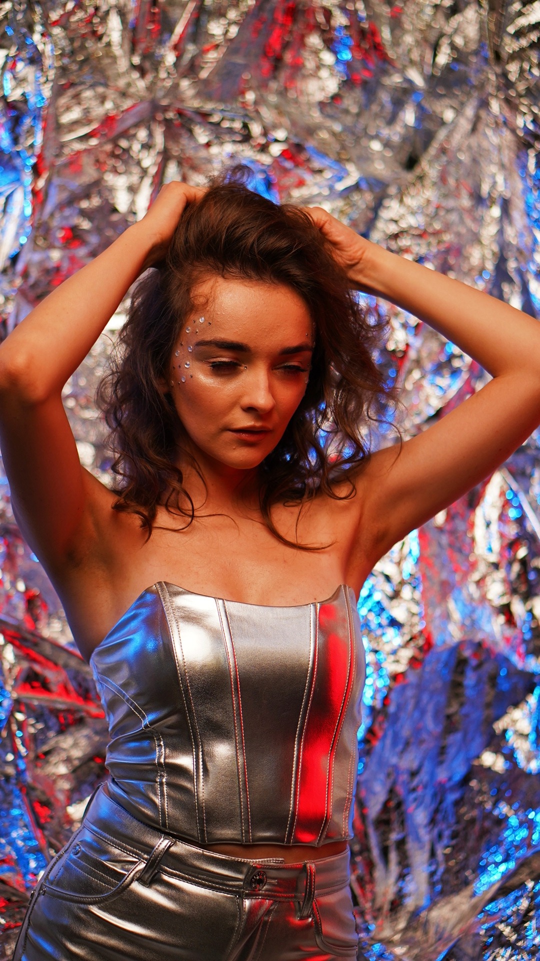 Portret kobiety w srebrnym topie i spodniach, z kryształkami na twarzy, pozującej na tle błyszczącej folii w odcieniach czerwieni i niebieskiego.