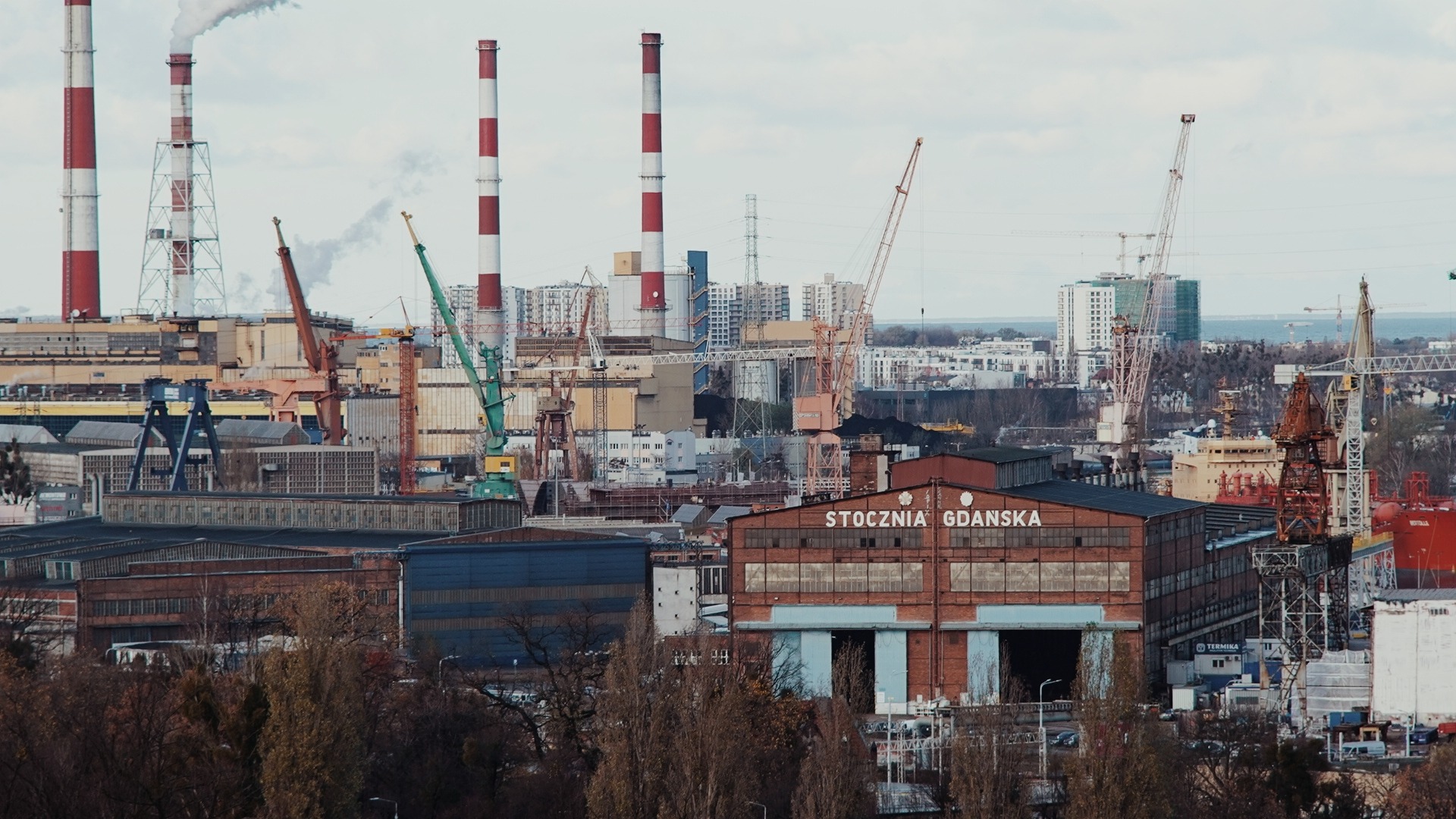 Panorama Stoczni Gdańskiej z kominami w biało-czerwone pasy, dźwigami i halami produkcyjnymi w tle zabudowa miejska.