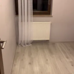 Pusta, jasna podłoga z paneli, biały grzejnik pod oknem z brązową ramą i zwiewna, biała firanka.