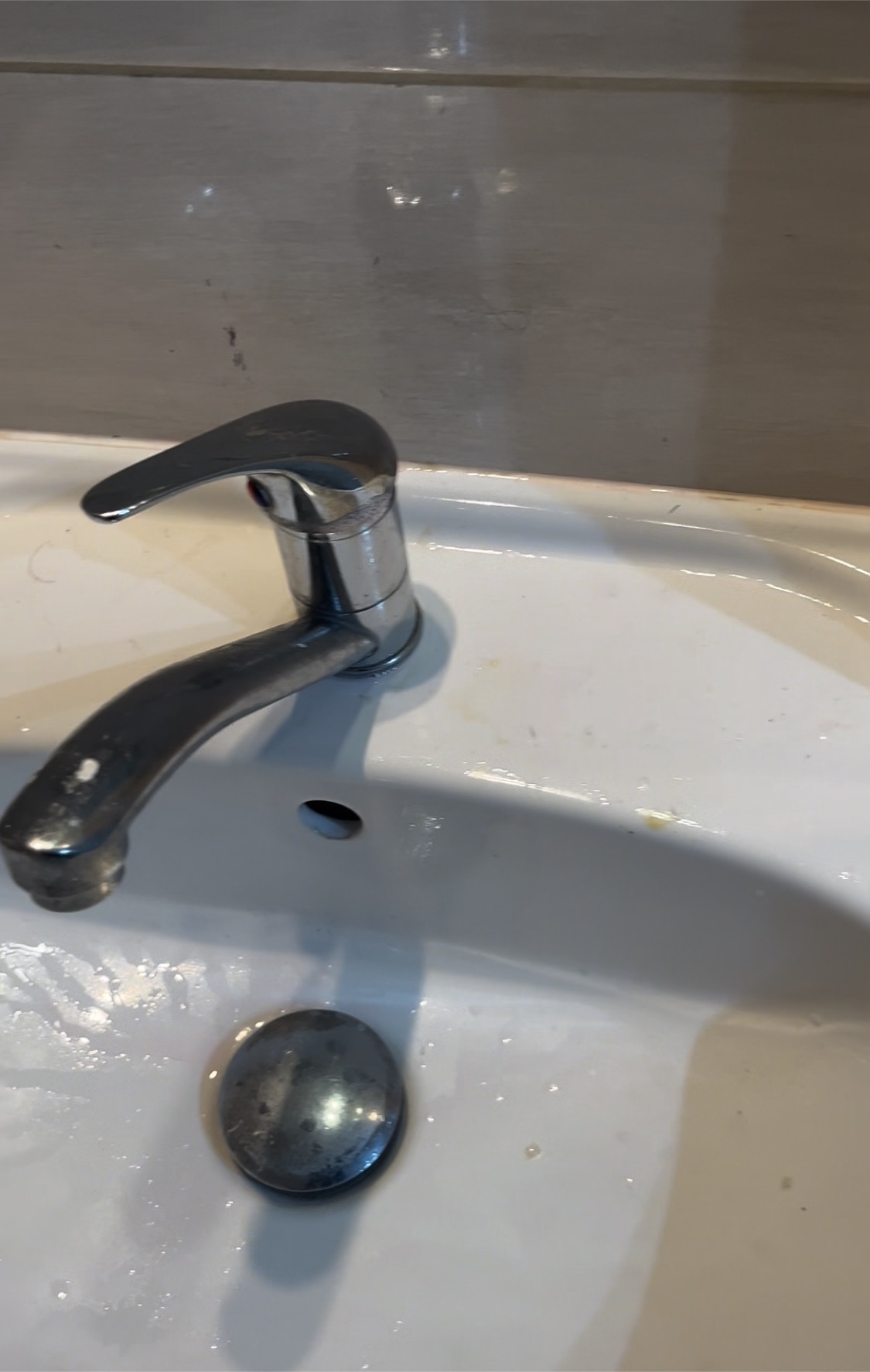 Biała umywalka z chromowaną baterią i zatkanym odpływem, widoczne ślady po zaschniętej wodzie i niewielkie zabrudzenia.