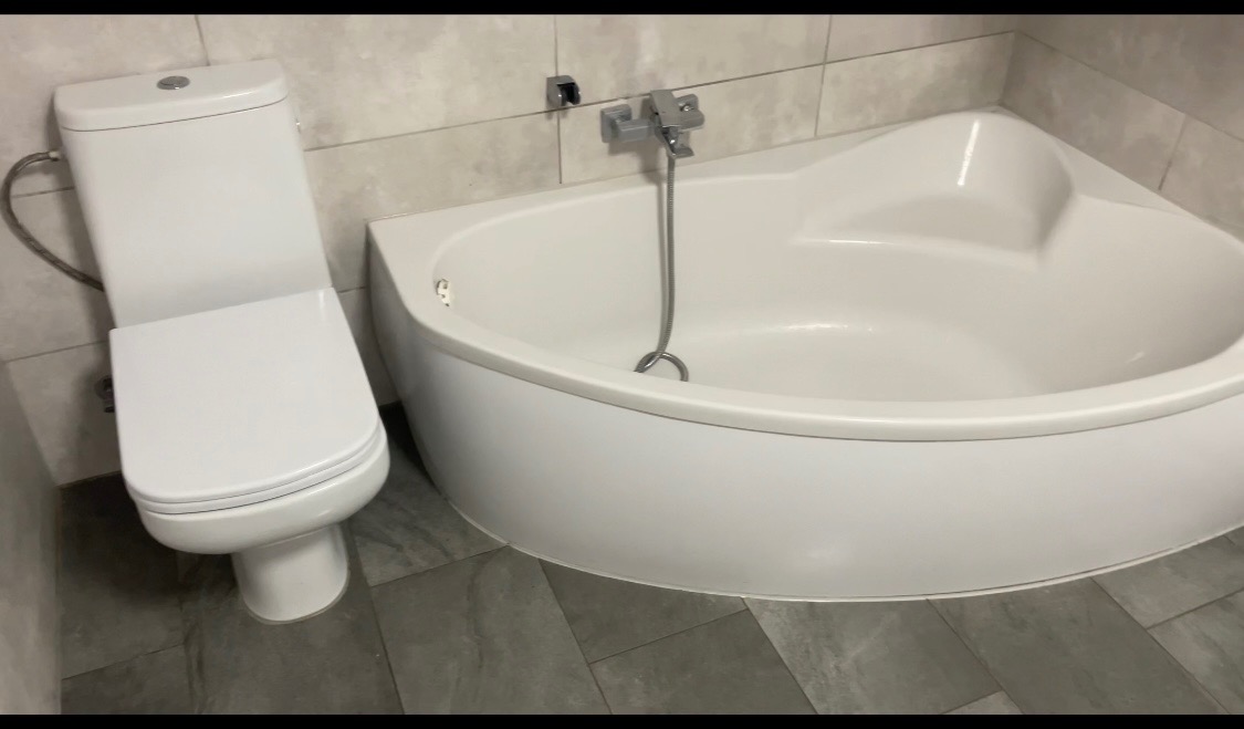 Biała narożna wanna z baterią i toaleta kompaktowa w łazience wykończonej szarymi płytkami.