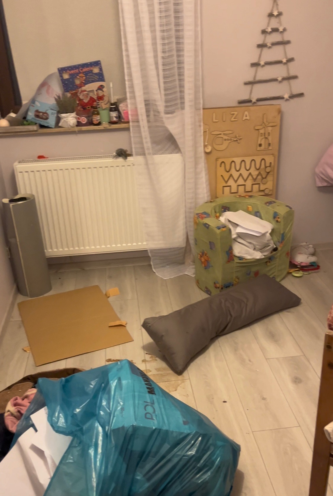 Pokój przed sprzątaniem: widoczny niebieski worek na śmieci wypełniony papierami, karton, poduszka na podłodze, dziecięcy fotel wypełniony papierami, zabawki na ścianie.