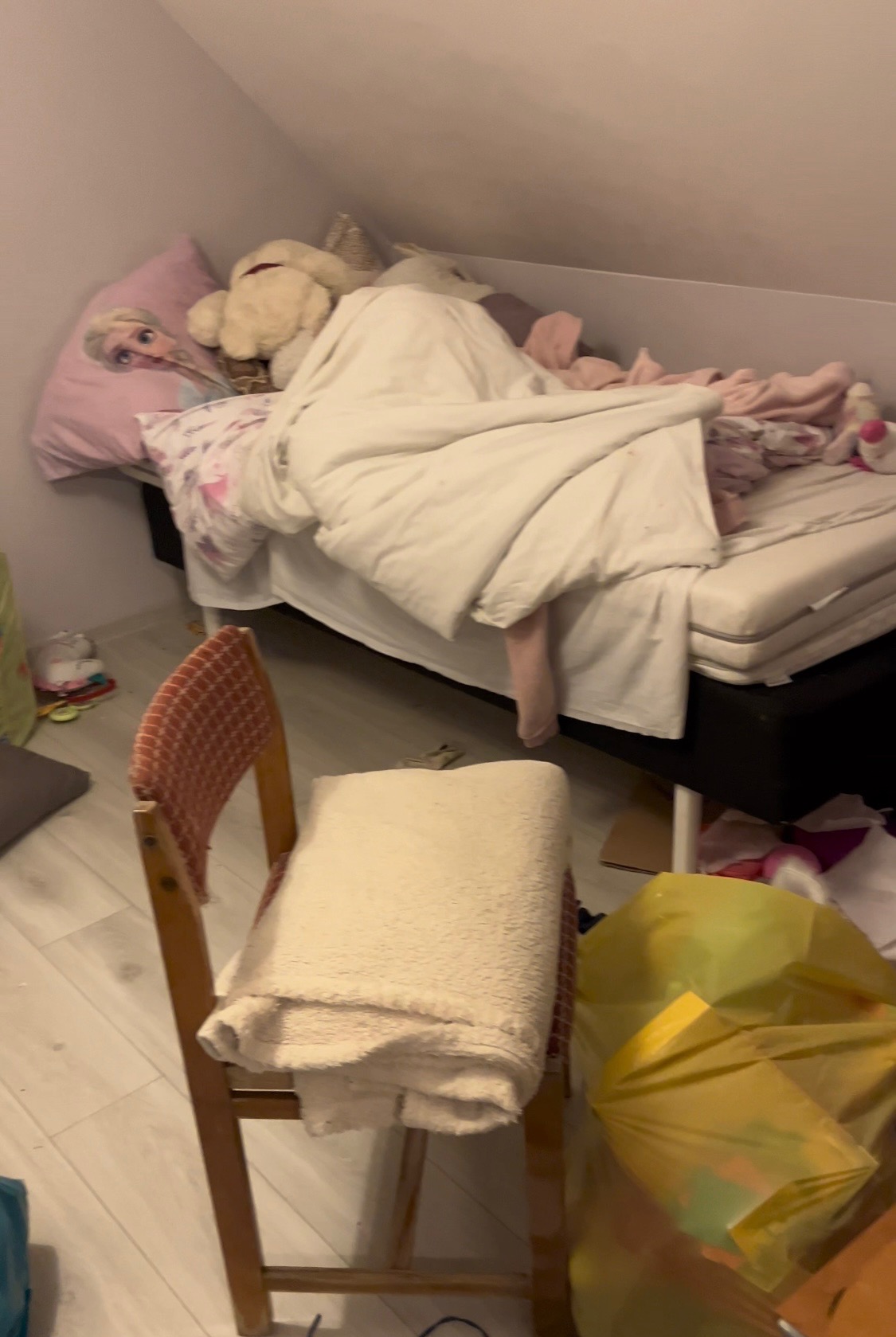 Zagracony pokój na poddaszu: niepościelone łóżko z poduszką z Elsą, pluszaki, krzesło z narzuconym pledem, worek na śmieci.