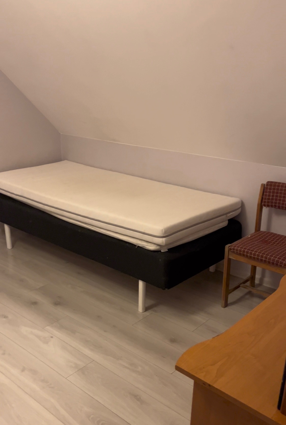 Stos materacy na czarnej ramie z białymi nogami w pokoju ze skośnym sufitem, obok krzesło z czerwono-białą kratą.