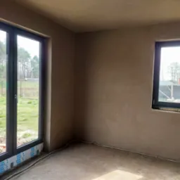 Wnętrze pokoju po tynkowaniu maszynowym, widoczne świeżo otynkowane ściany, okna PCV z folią ochronną VEKA, oraz wystające kable elektryczne przy podłodze.