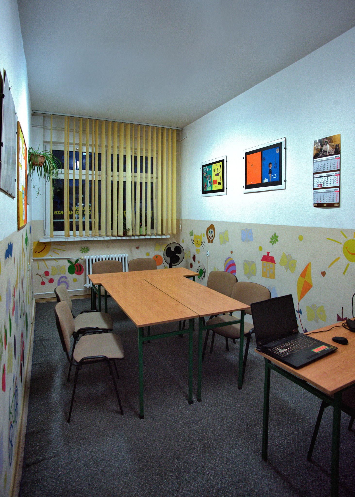 Sala lekcyjna z drewnianymi stołami i krzesłami, ściany ozdobione dziecięcymi rysunkami, na biurku otwarty laptop, w tle okno z żaluzjami.