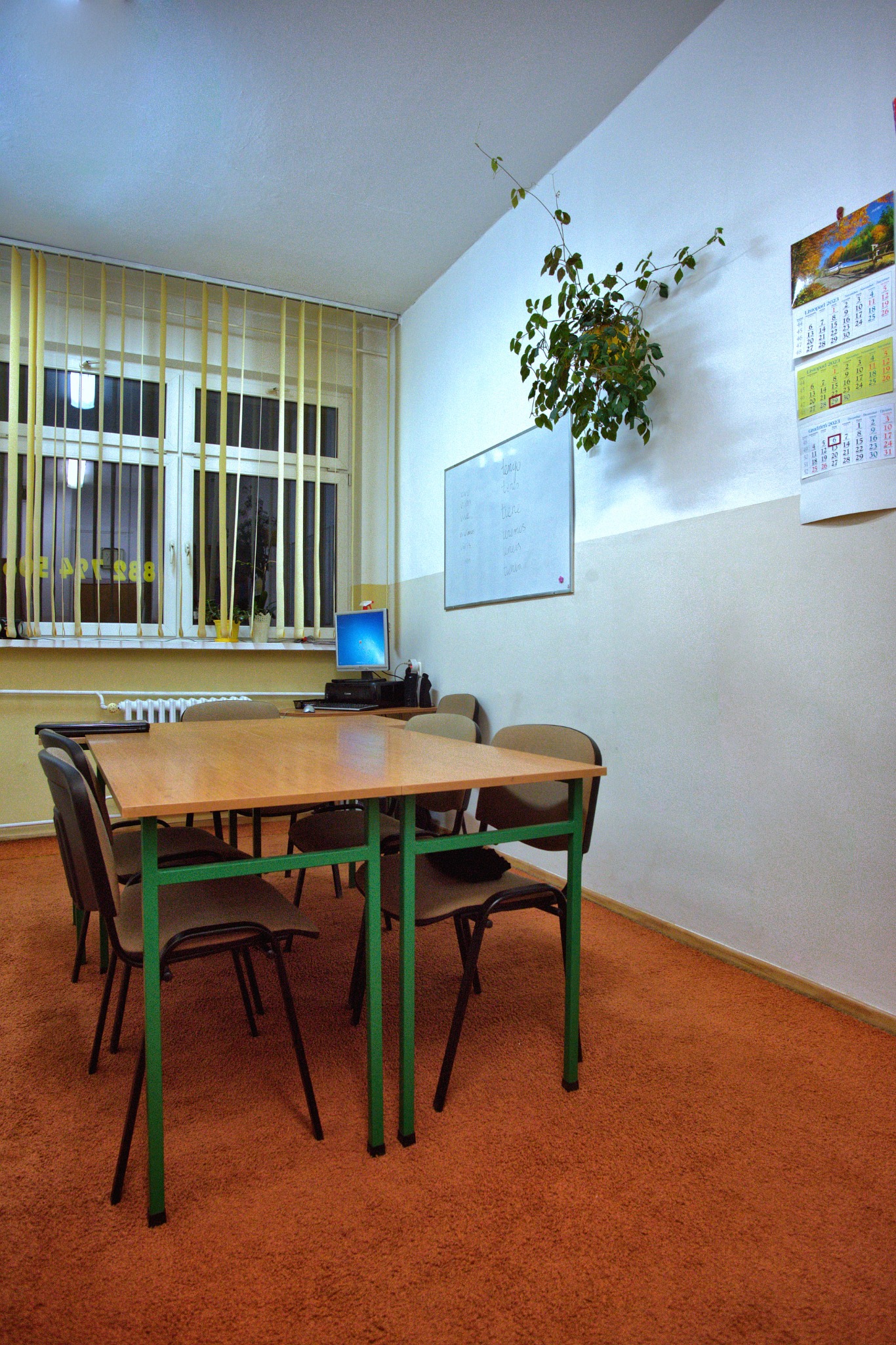 Sala lekcyjna z drewnianymi stołami z zielonymi nogami, krzesłami, tablicą z zapiskami, kalendarzem na ścianie i rośliną wiszącą w rogu.