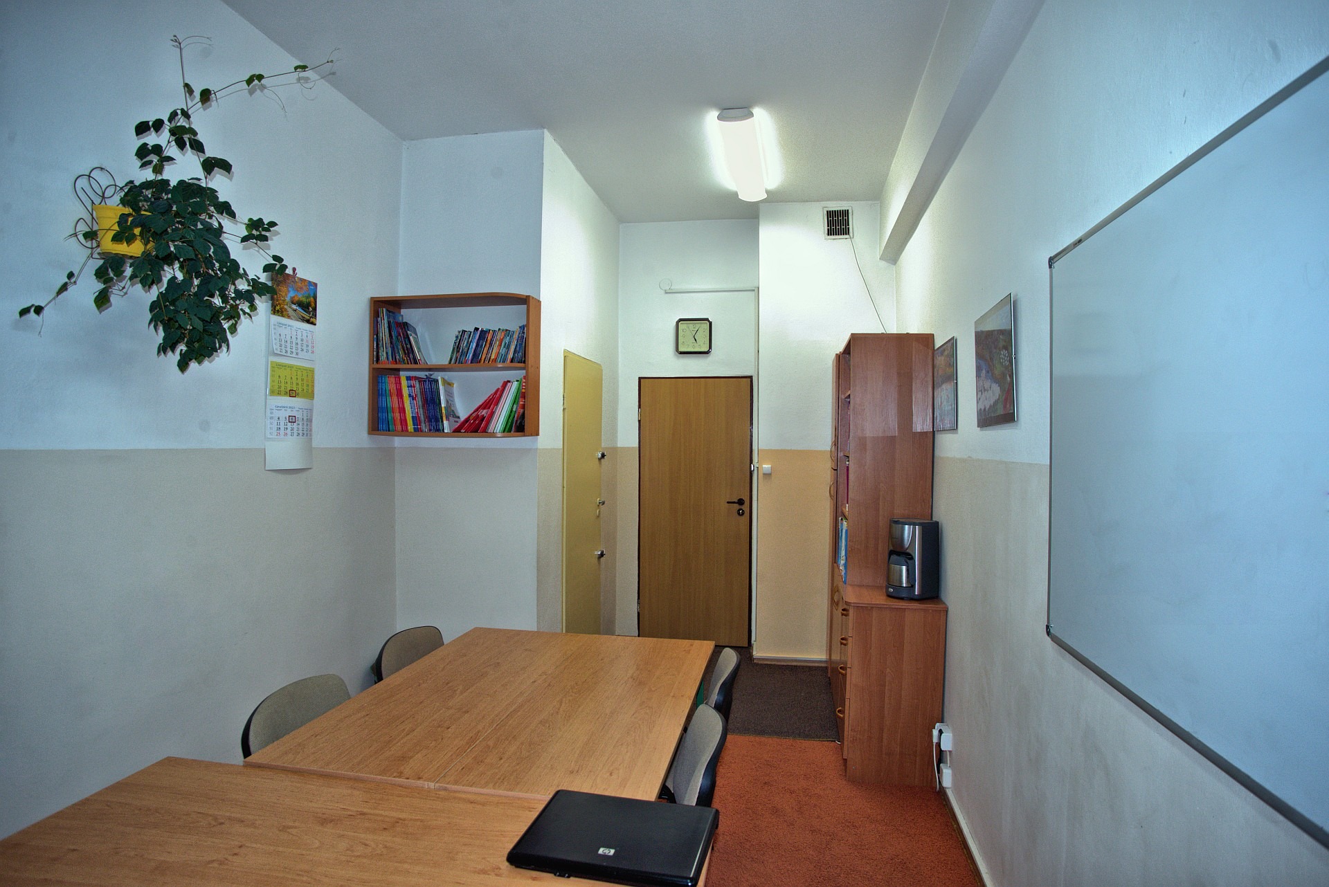 Wnętrze klasy językowej z drewnianymi ławkami, tablicą, regałem z książkami, kwiatem doniczkowym i laptopem na blacie.