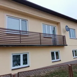 Dwupiętrowy dom z beżową elewacją, brązową balustradą balkonową z poziomych listew i satelitą na ścianie, widziany zza metalowej siatki ogrodzeniowej.