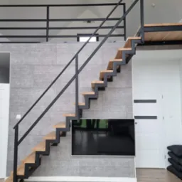 Nowoczesne schody wewnętrzne z drewnianymi stopniami i czarną metalową konstrukcją, zamontowane przy szarej ścianie z imitacją betonu, widoczne drzwi i telewizor.
