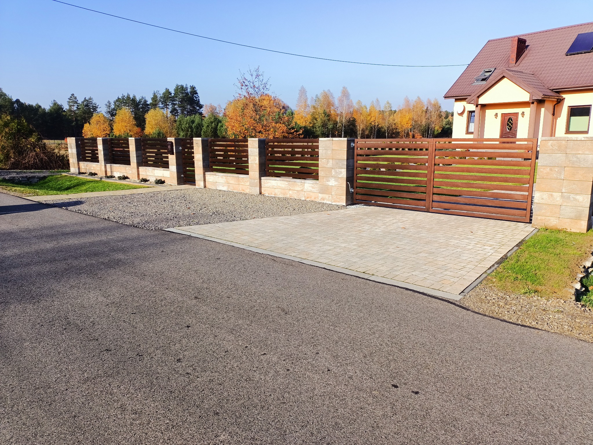 Nowoczesne ogrodzenie poziome z brązowych paneli i beżowych słupków z kostki, przesuwna brama wjazdowa, podjazd wyłożony kostką i żwirem, jesienne drzewa w tle.