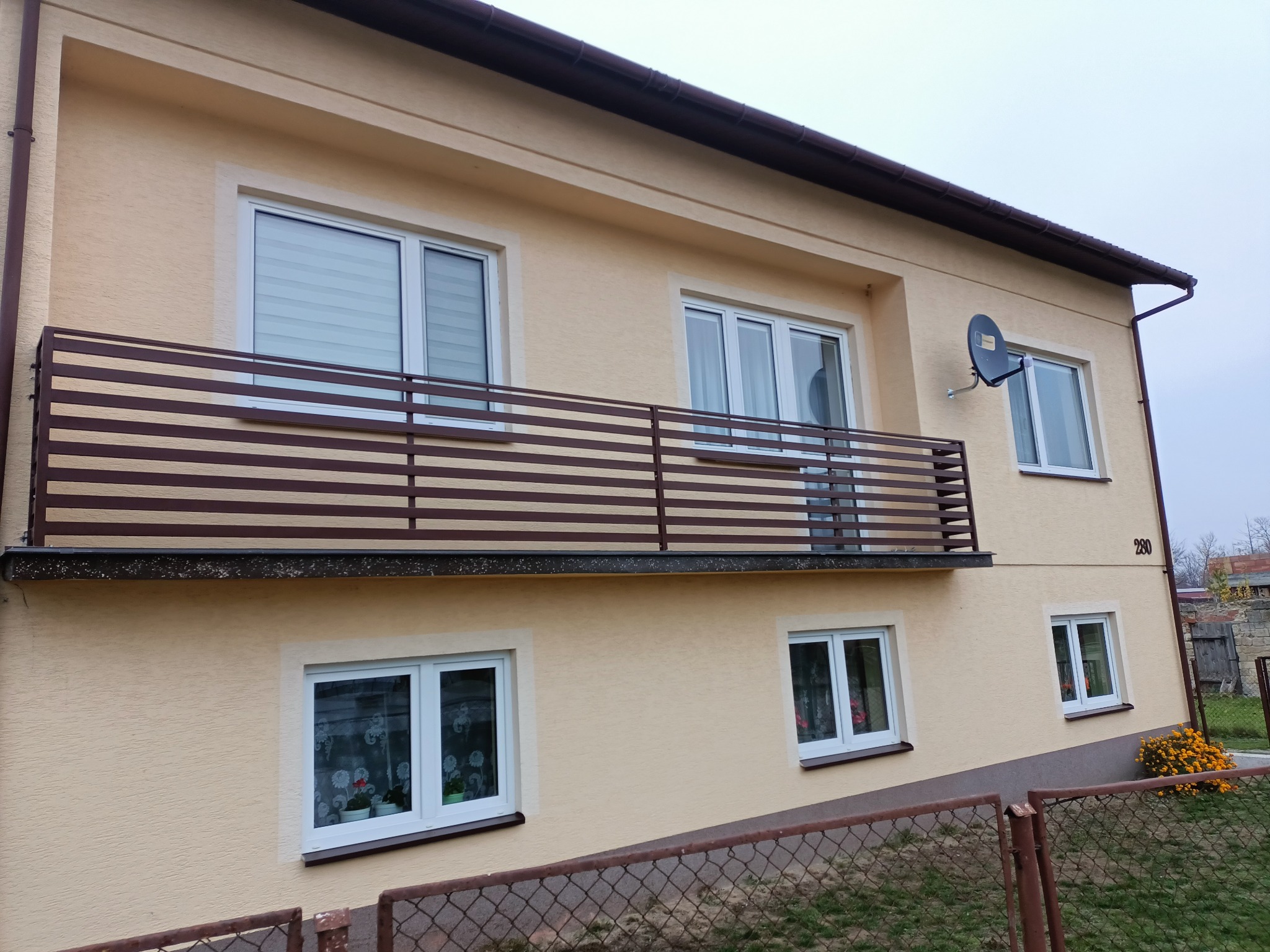 Dwupiętrowy dom z beżową elewacją, brązową balustradą balkonową z poziomych listew i satelitą na ścianie, widziany zza metalowej siatki ogrodzeniowej.