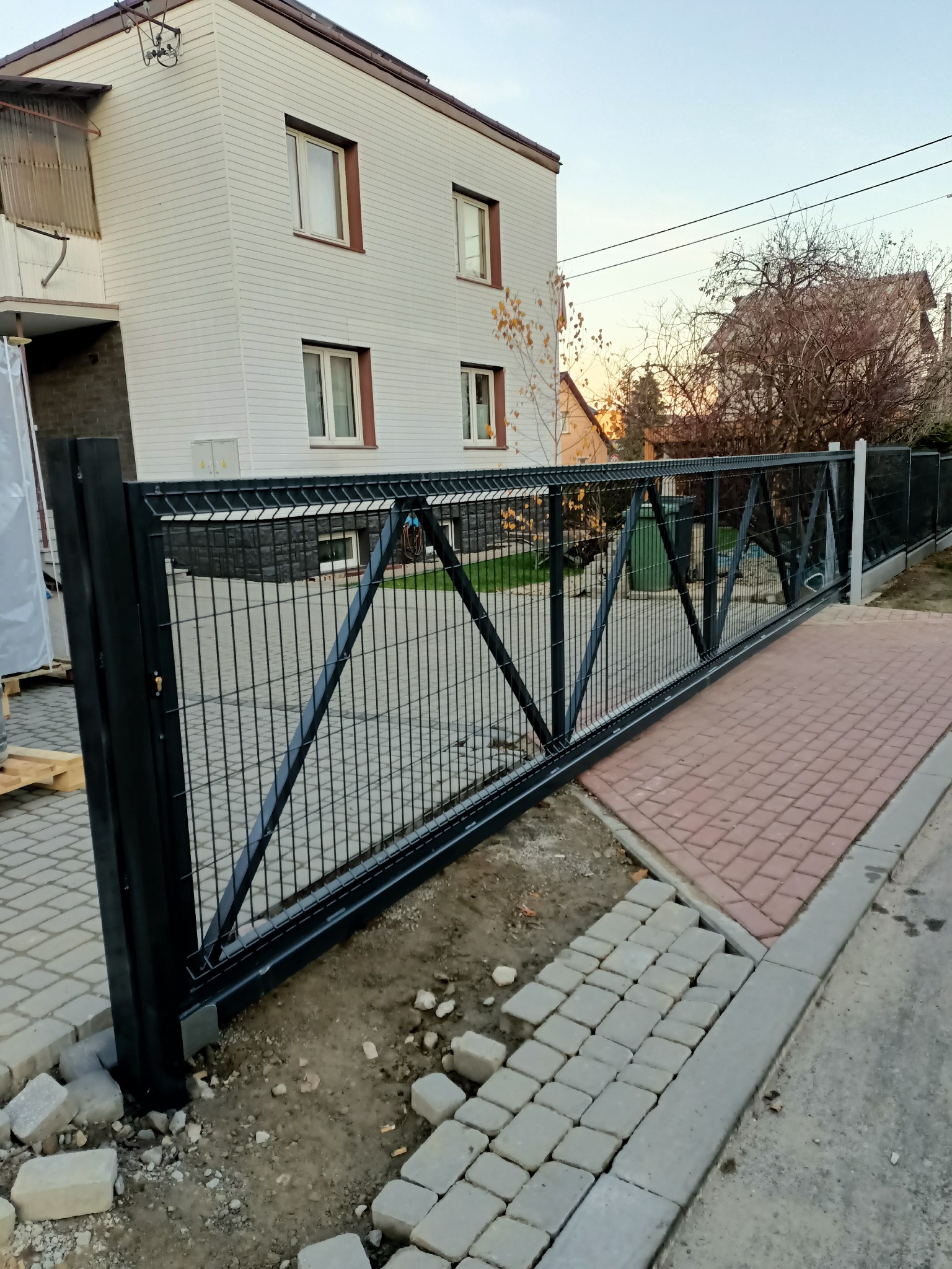 Nowoczesna, przesuwna brama panelowa w kolorze grafitowym, z widocznymi elementami konstrukcyjnymi i niedokończonym układaniem kostki brukowej wokół, na tle domu z jasną elewacją.