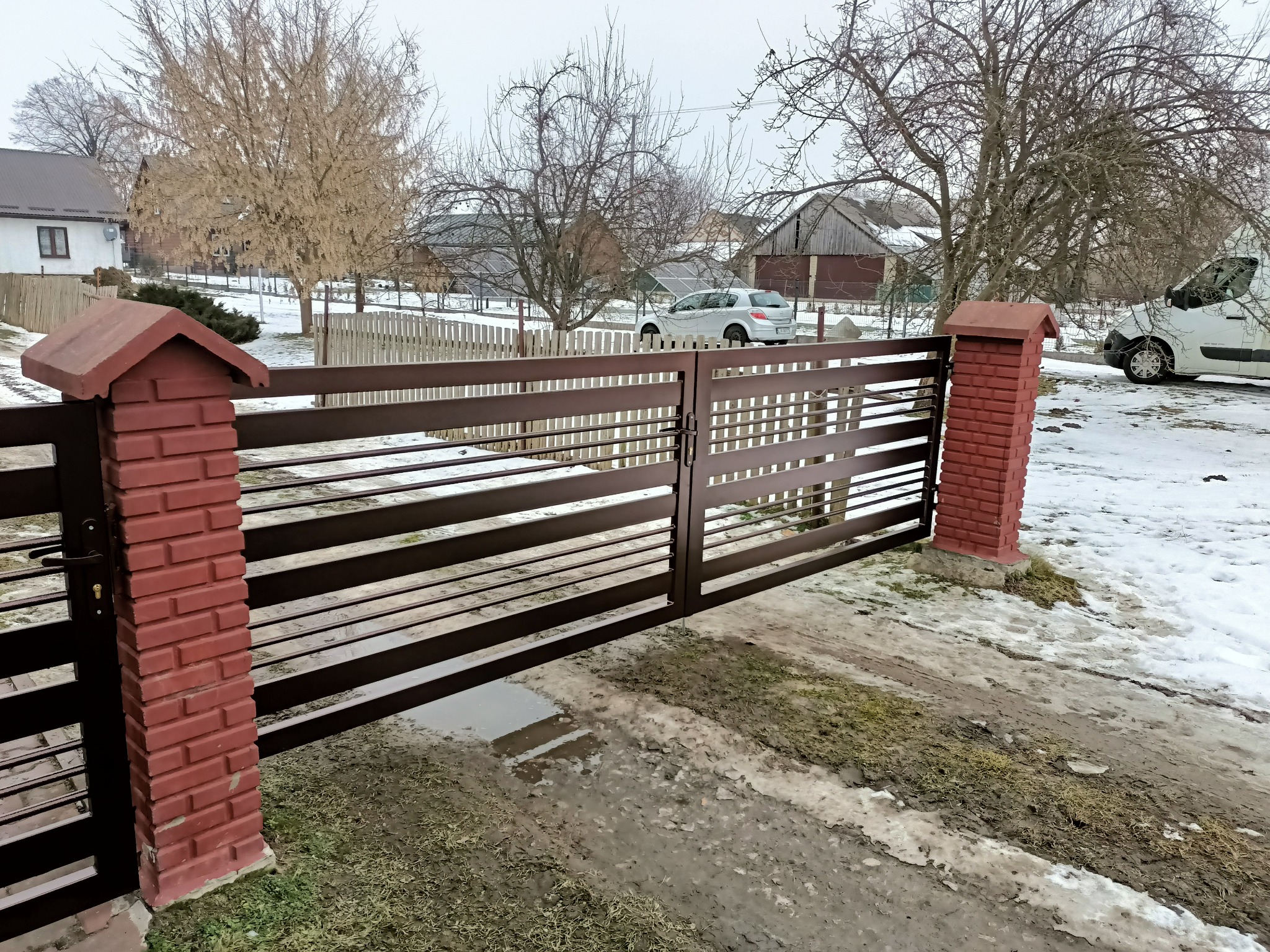 Nowoczesna brama wjazdowa z poziomych profili w kolorze brązowym, osadzona na ceglanych słupkach z daszkami, widok na posesję z samochodem osobowym i zabudowaniami w zimowej scenerii.