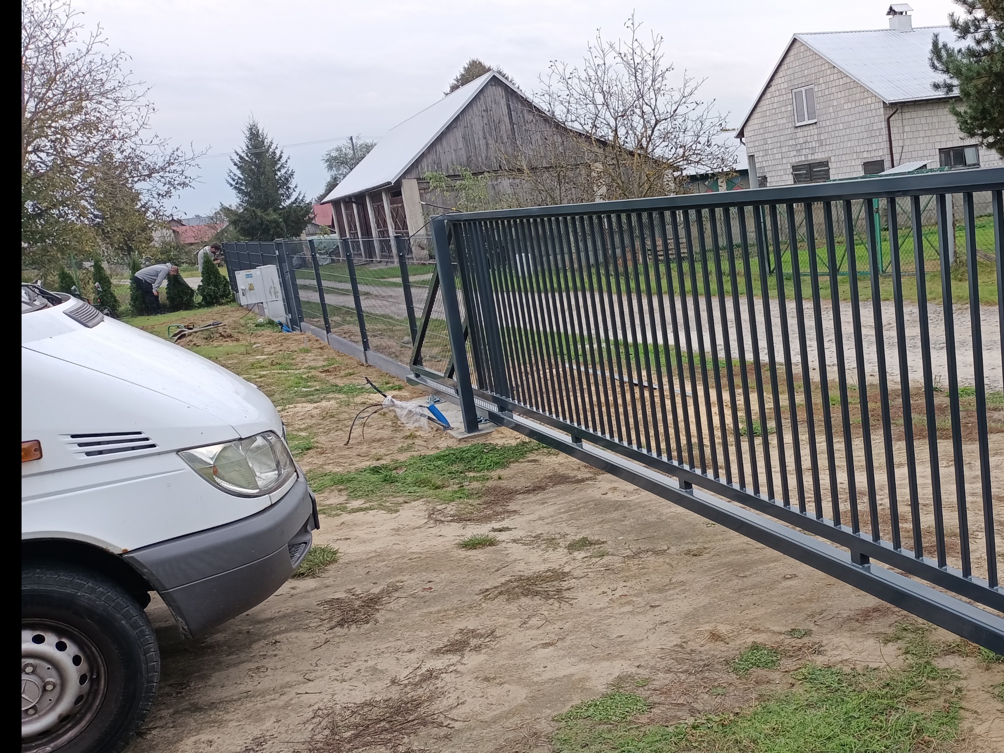 Nowoczesna, przesuwna brama wjazdowa z pionowych profili, częściowo otwarta, widoczny samochód dostawczy i pracownicy w tle, teren budowy.