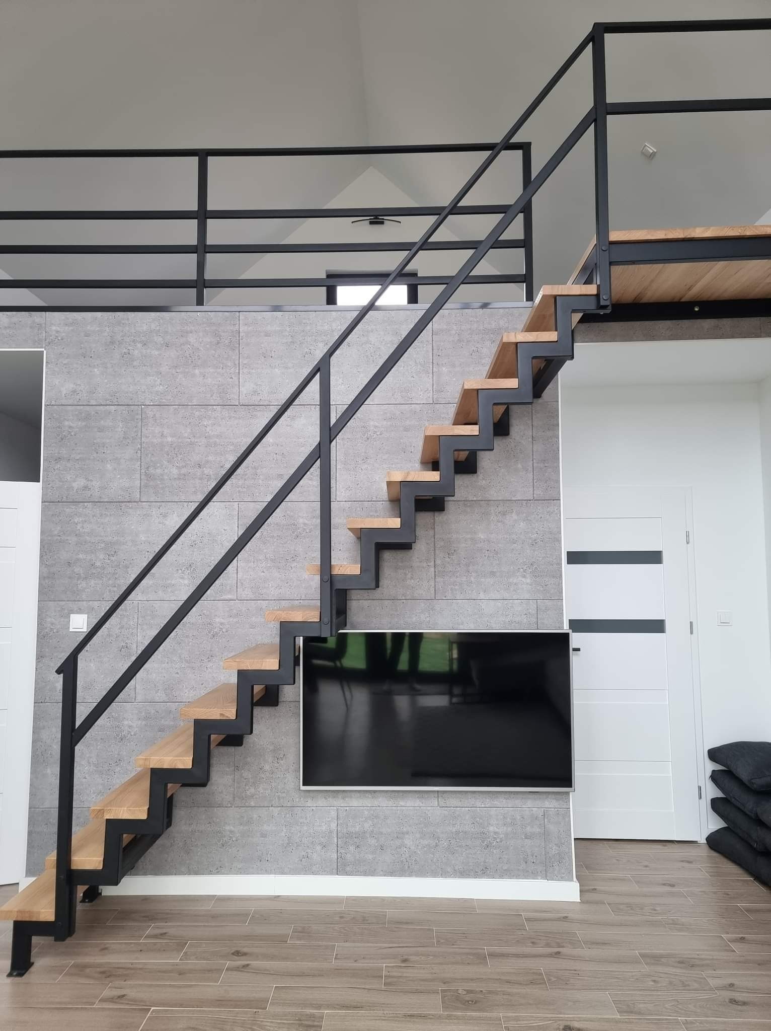 Nowoczesne schody wewnętrzne z drewnianymi stopniami i czarną metalową konstrukcją, zamontowane przy szarej ścianie z imitacją betonu, widoczne drzwi i telewizor.