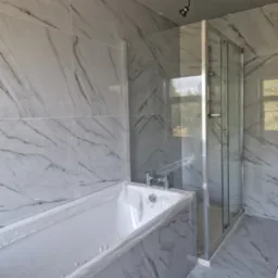 Niedokończona łazienka z marmurowymi płytkami, osłoniętą folią wanną i kabiną prysznicową, widoczne niedociągnięcia w suficie.