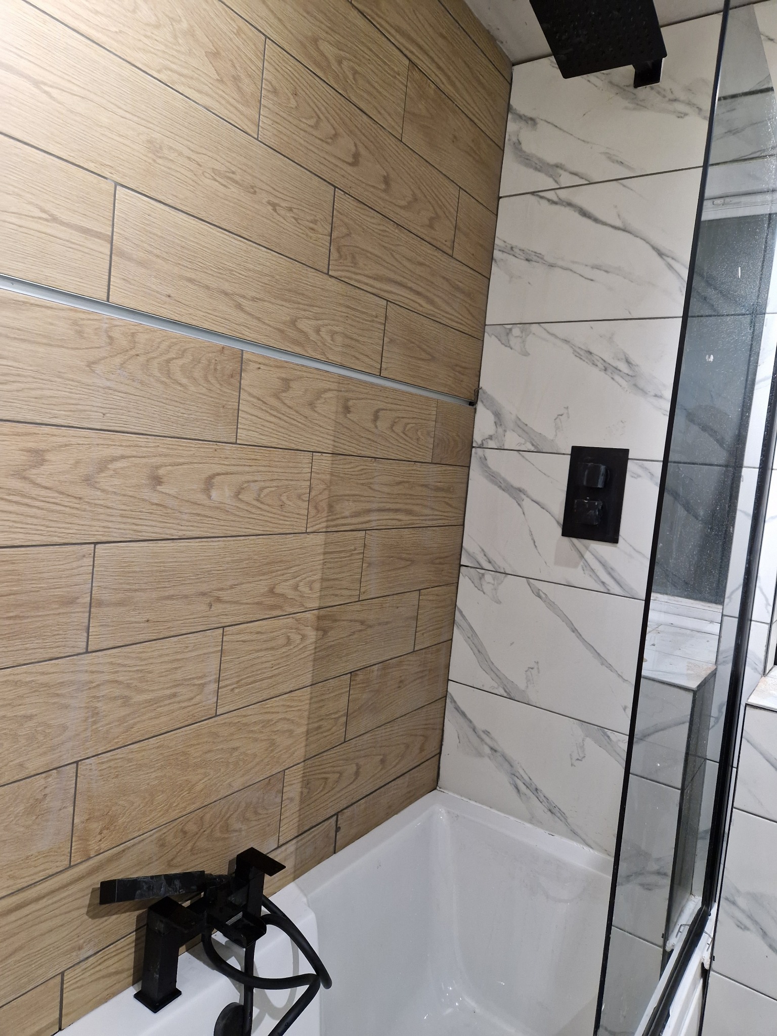 Nowoczesna łazienka z drewnopodobnymi płytkami na ścianie, białą wanną i czarną armaturą, oddzielona szklaną ścianką prysznicową, z marmurowymi płytkami po prawej stronie.