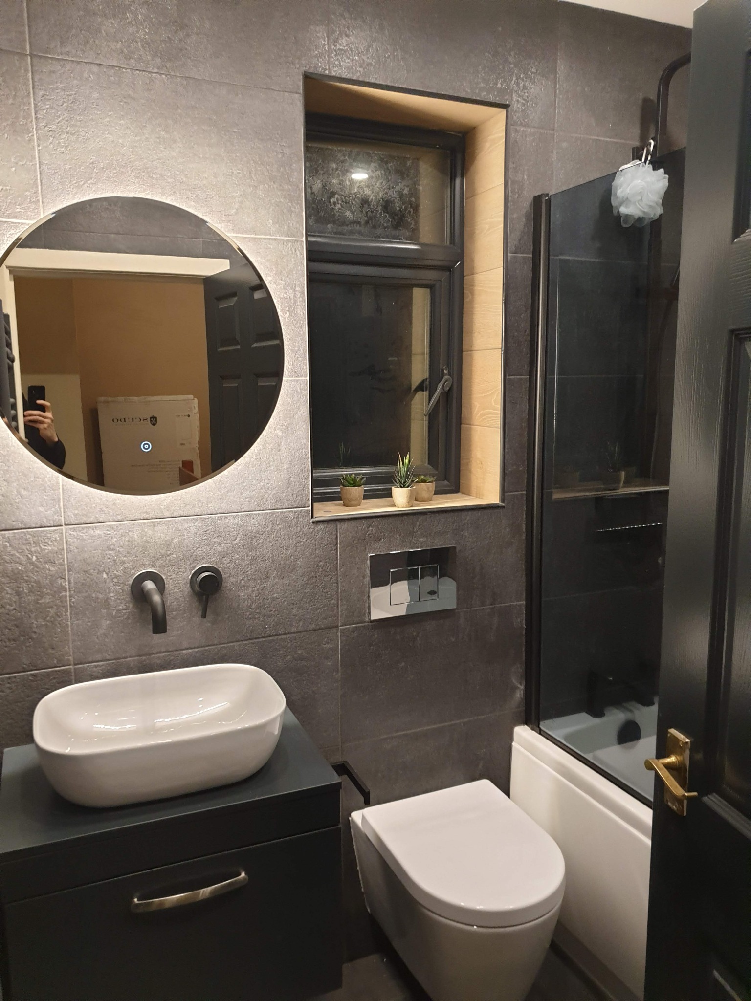 Nowoczesna łazienka z szarymi płytkami, owalnym lustrem z podświetleniem LED, białą umywalką nablatową, czarną armaturą, oknem z roślinami i kabiną prysznicową z czarnymi ramami.