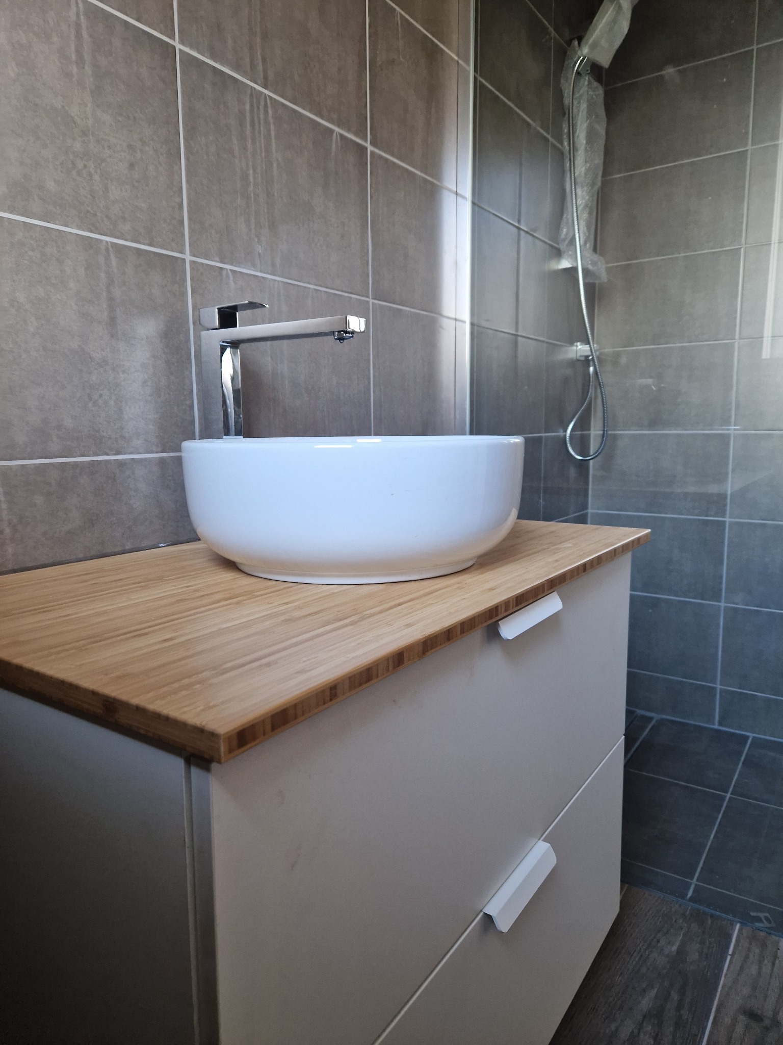 Nowoczesna łazienka z umywalką nablatową, drewnianym blatem i szarą glazurą, bateria umywalkowa chromowana, kabina prysznicowa w folii ochronnej.