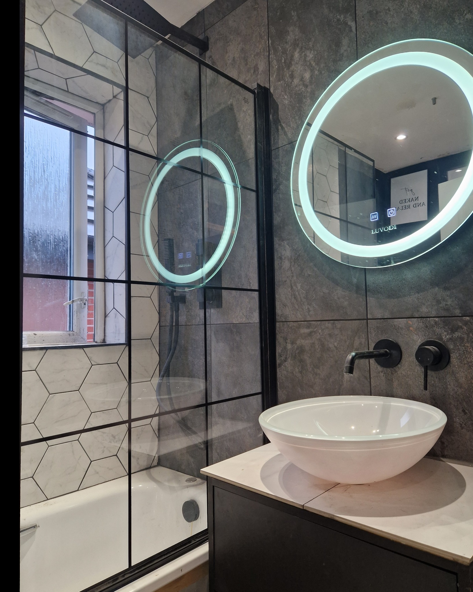 Nowoczesna łazienka z kabiną prysznicową oddzieloną czarną ramą, okrągłe lustro z podświetleniem LED, umywalka nablatowa na czarnej szafce, heksagonalne płytki na ścianie przy wannie.