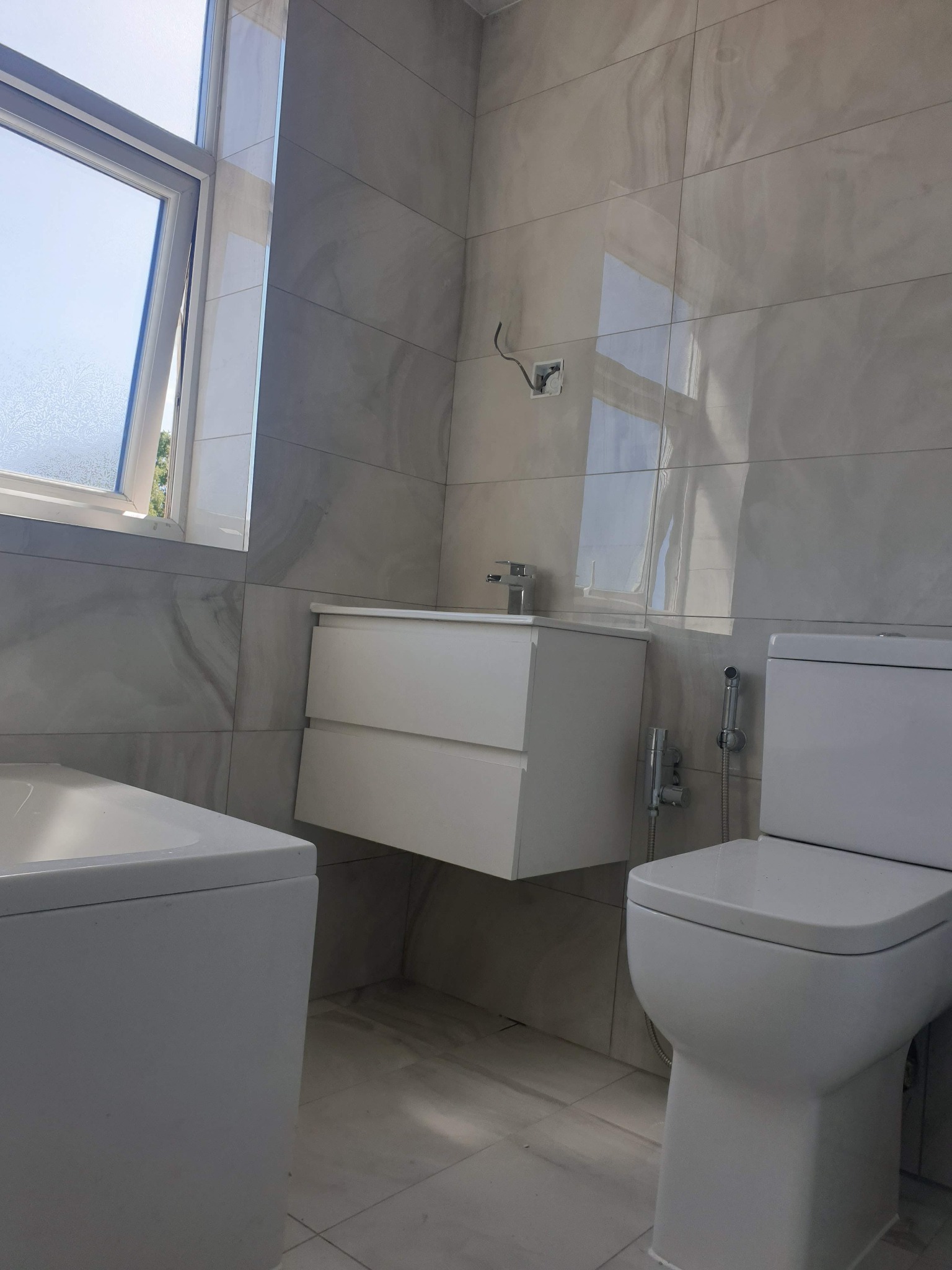 Jasna łazienka z wanną, podwieszaną szafką z umywalką i toaletą; szare płytki imitujące marmur na ścianach, widoczne niedokończone podłączenie elektryczne.