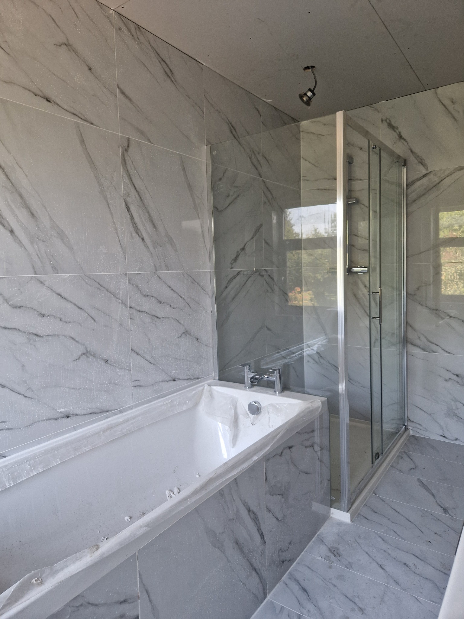 Niedokończona łazienka z marmurowymi płytkami, osłoniętą folią wanną i kabiną prysznicową, widoczne niedociągnięcia w suficie.