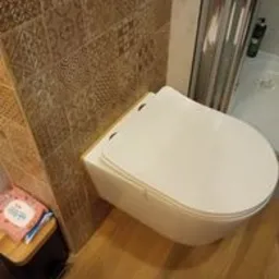 Wisząca toaleta w nowoczesnej łazience z brązowymi płytkami o geometrycznym wzorze, drewnopodobna podłoga, widoczny fragment kabiny prysznicowej i kosmetyki na małym stoliczku.