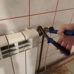 Hydraulik dokręca złączkę przy białym grzejniku panelowym za pomocą klucza nastawnego z niebieskimi uchwytami. Widoczne płytki ceramiczne z czerwoną fugą w tle.