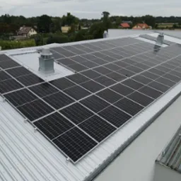 Instalacja paneli słonecznych na dachu przemysłowym z szarej blachy trapezowej, widoczne kominki wentylacyjne, w tle zabudowa jednorodzinna.