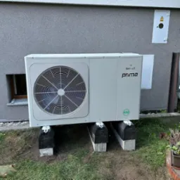 Zewnętrzna jednostka pompy ciepła Galmet Prima R32 zamontowana na betonowych bloczkach z gumowymi podkładkami, ustawiona przy szarej elewacji budynku z małym oknem piwnicznym.