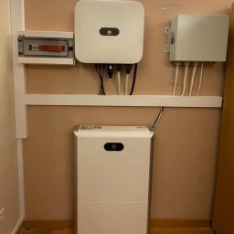 ECOFAB - Biały magazyn energii z logo Huawei, skrzynka elektryczna i inwerter zamontowane na ścianie w pomieszczeniu.