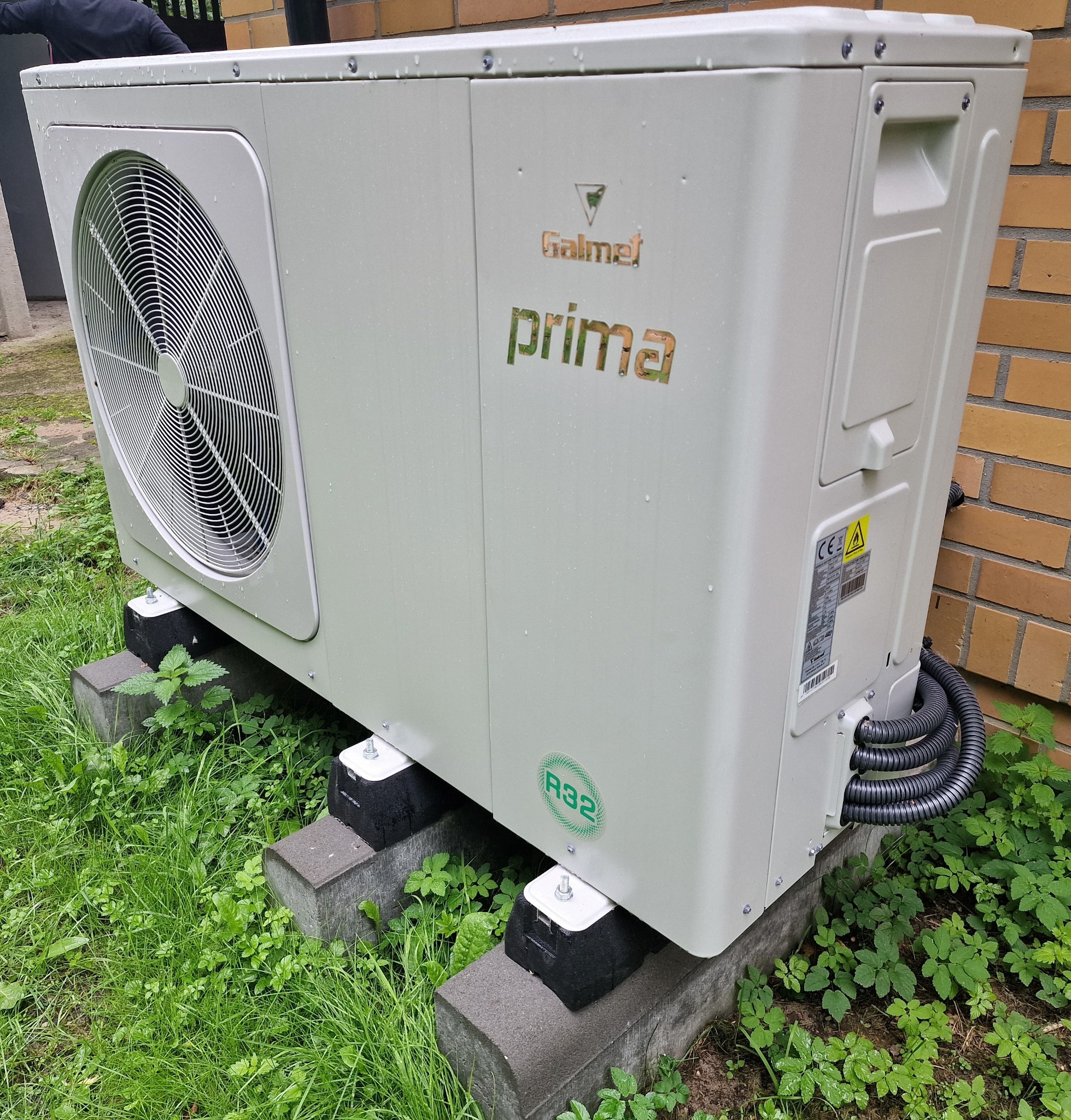 Biała jednostka zewnętrzna z wentylatorem, marki Galmet Prima, zamontowana na betonowych bloczkach w trawie, widoczne przewody i fragment ceglanej ściany.