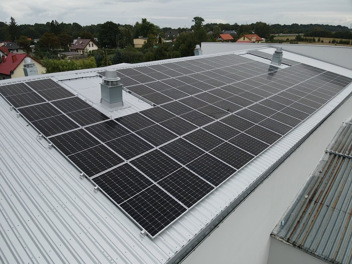 Instalacja paneli słonecznych na dachu przemysłowym z szarej blachy trapezowej, widoczne kominki wentylacyjne, w tle zabudowa jednorodzinna.