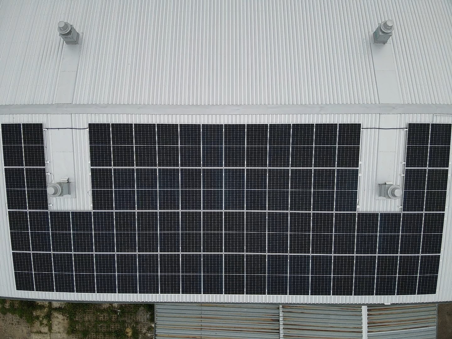 Widok z góry na instalację fotowoltaiczną na blaszanym dachu budynku przemysłowego z czterema wentylatorami dachowymi.