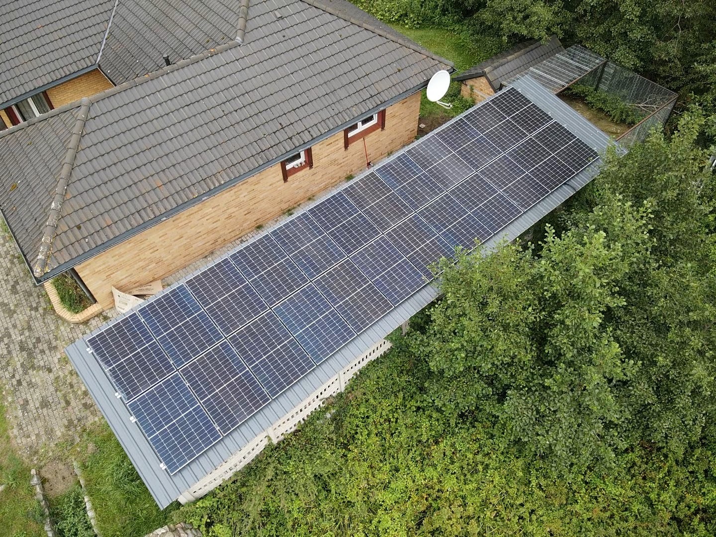 Widok z góry na panele słoneczne zamontowane na płaskim dachu budynku gospodarczego, obok domu jednorodzinnego z brązową dachówką, otoczonego zielenią.