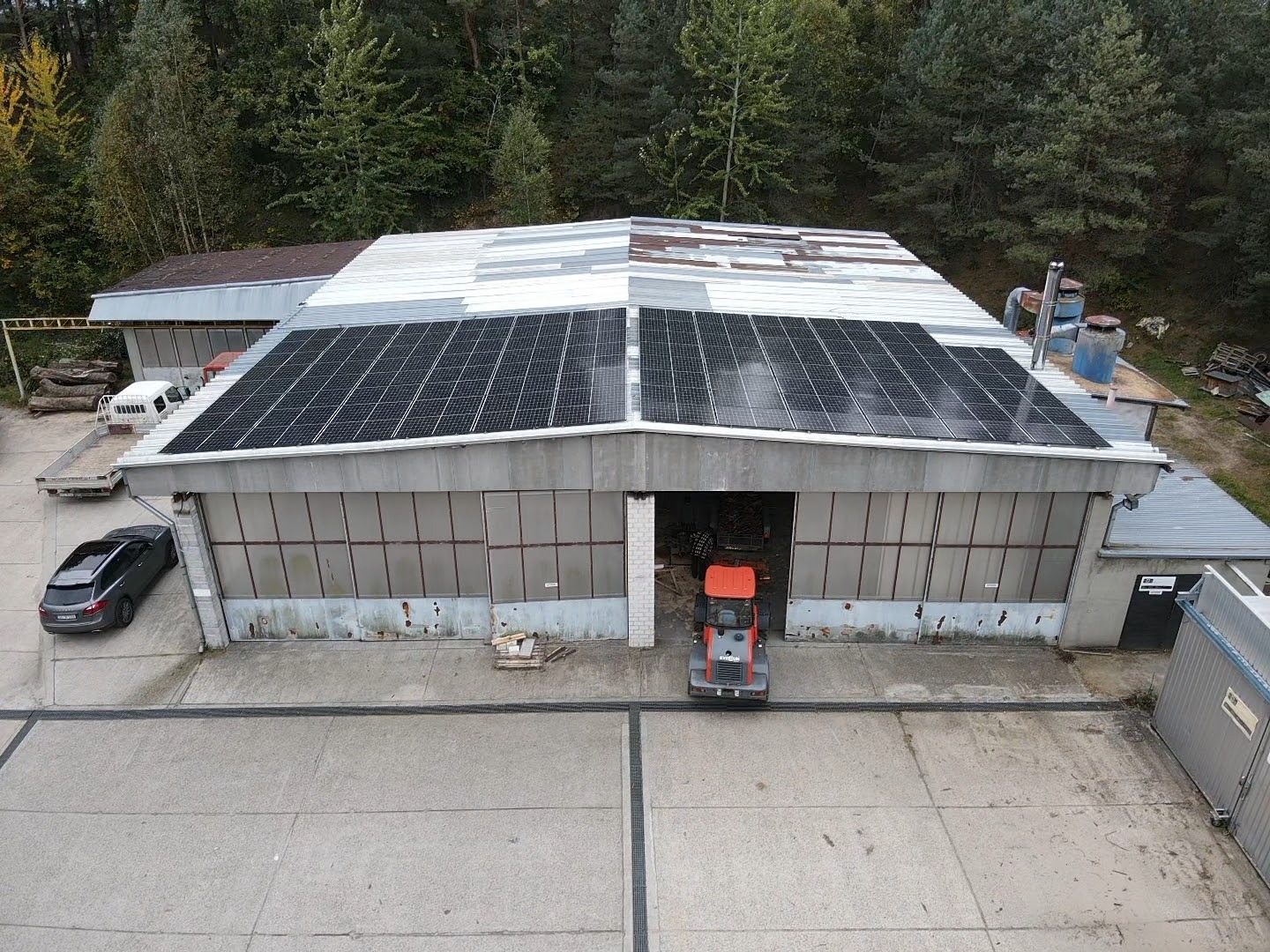 Hala przemysłowa z panelami fotowoltaicznymi na dachu, widok z góry, obok zaparkowany samochód, wewnątrz hali widoczny wózek widłowy