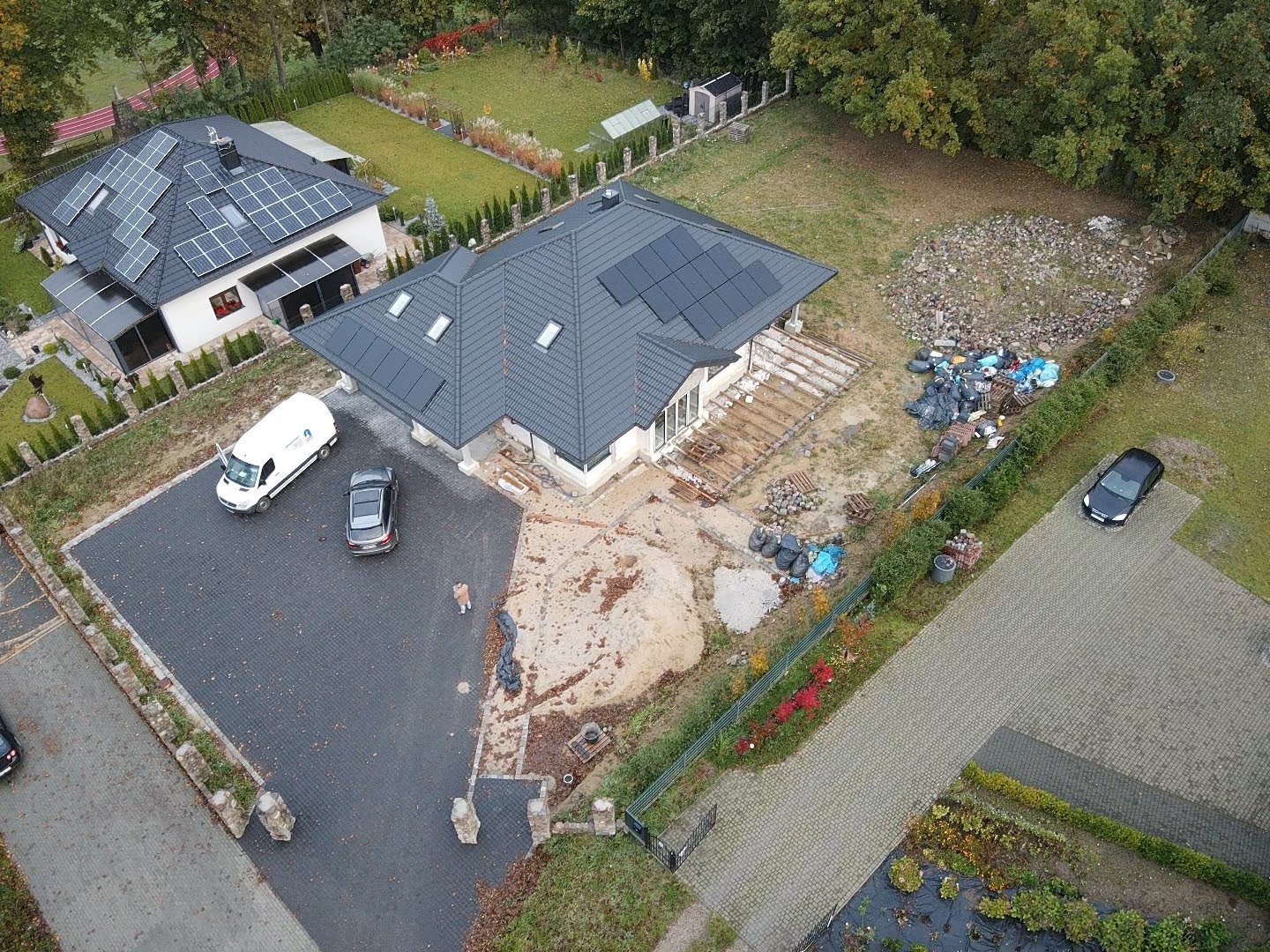 Widok z lotu ptaka na dwa domy jednorodzinne z panelami słonecznymi na dachach, biały bus serwisowy zaparkowany na podjeździe, teren wokół w trakcie prac wykończeniowych.