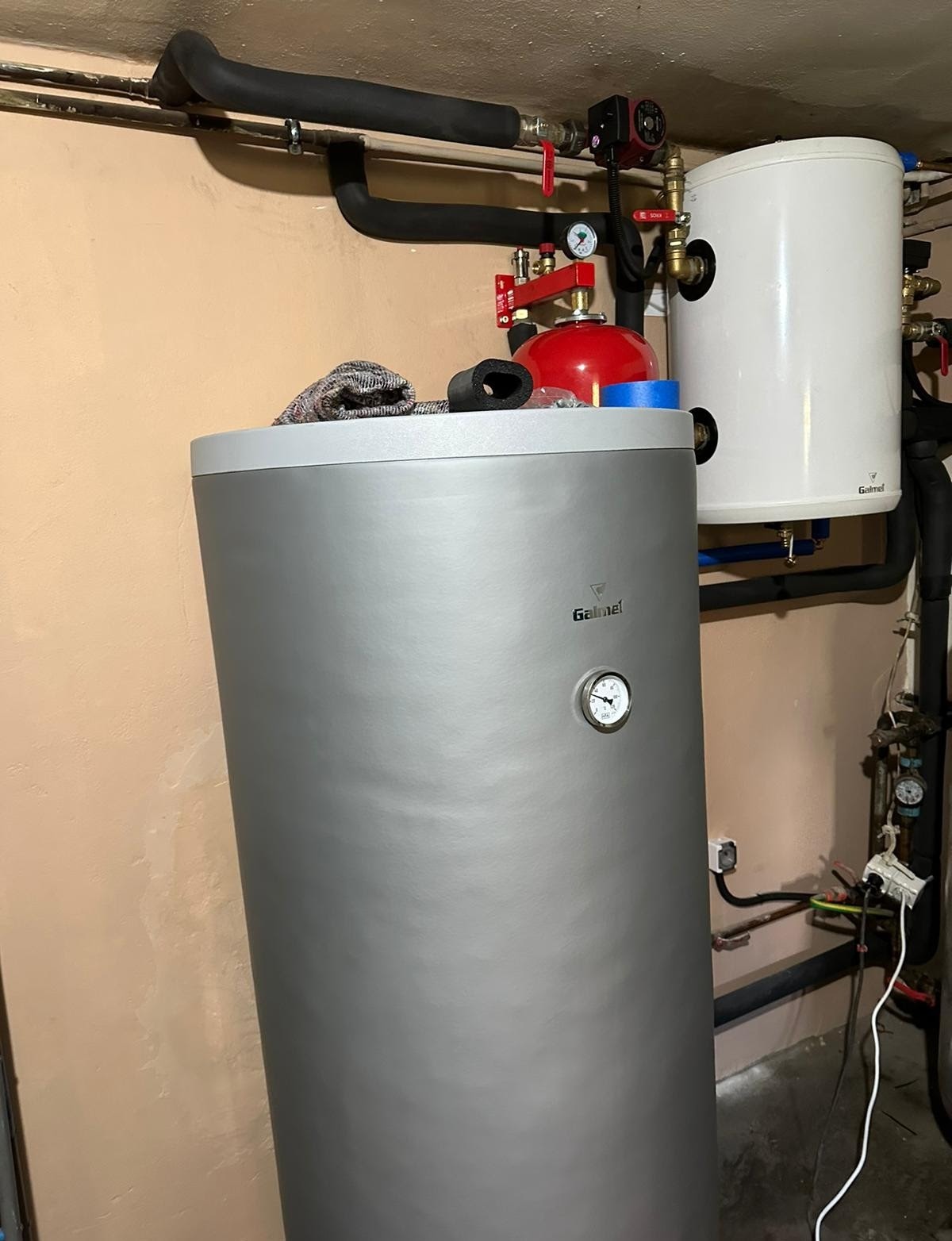 Instalacja z dwoma zbiornikami, szarym i białym, z widocznymi rurami, zaworami i manometrami, zamontowana przy beżowej ścianie, w pomieszczeniu o surowym wykończeniu.