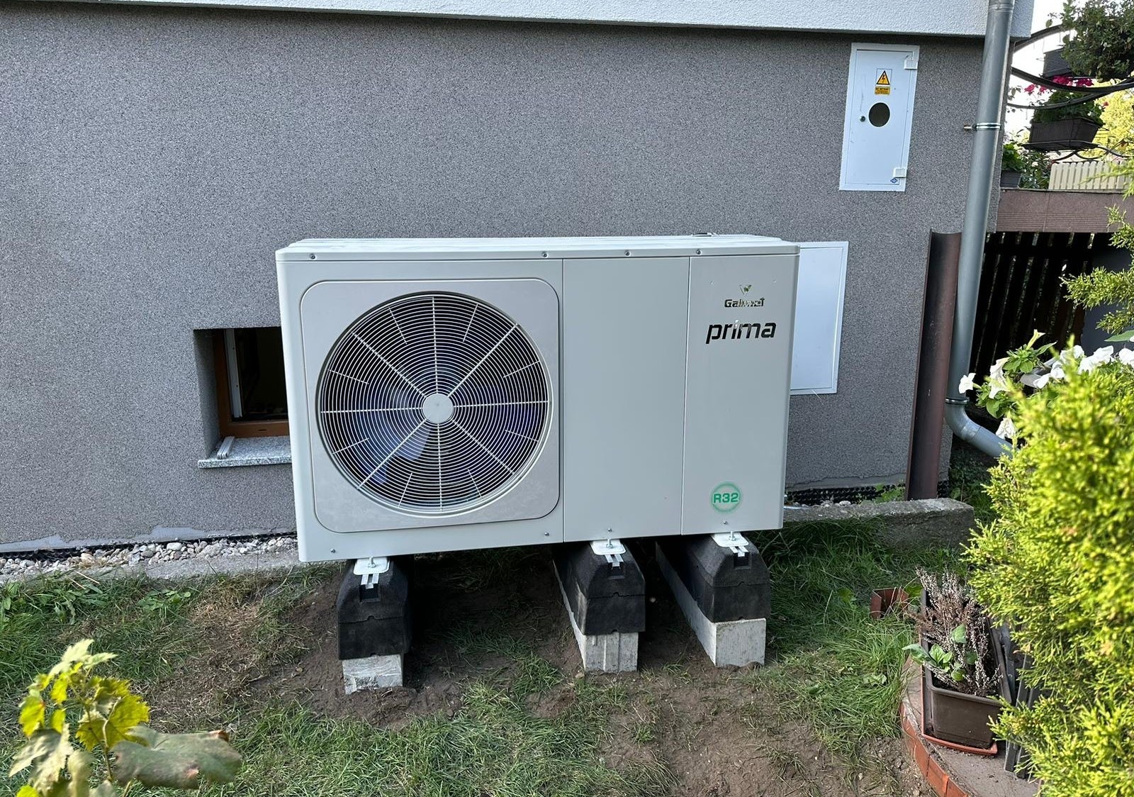 Zewnętrzna jednostka pompy ciepła Galmet Prima R32 zamontowana na betonowych bloczkach z gumowymi podkładkami, ustawiona przy szarej elewacji budynku z małym oknem piwnicznym.