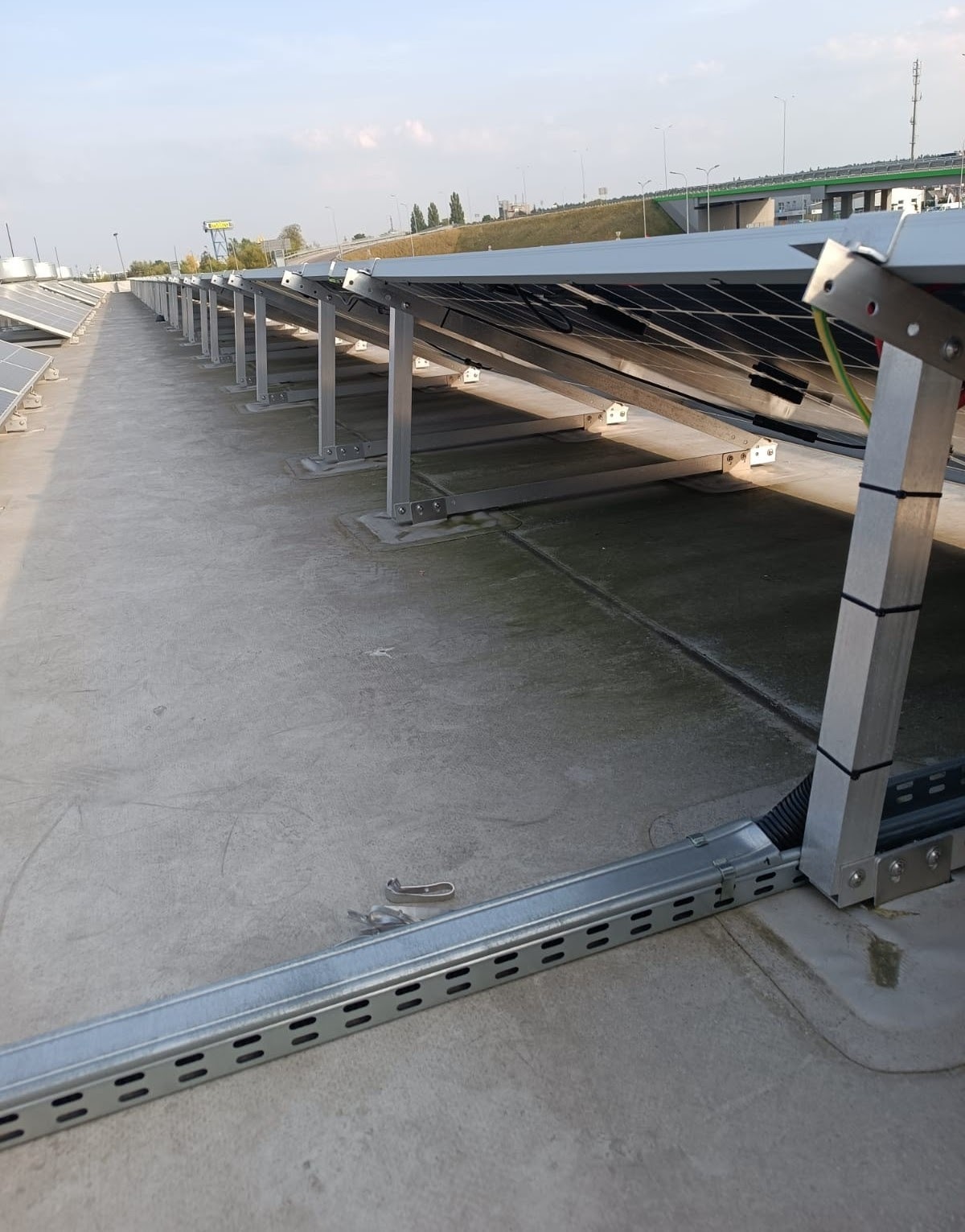 Rząd paneli słonecznych zamontowanych na płaskim dachu, widoczne elementy montażowe i kable w peszlu, w tle wiadukt i niebo.