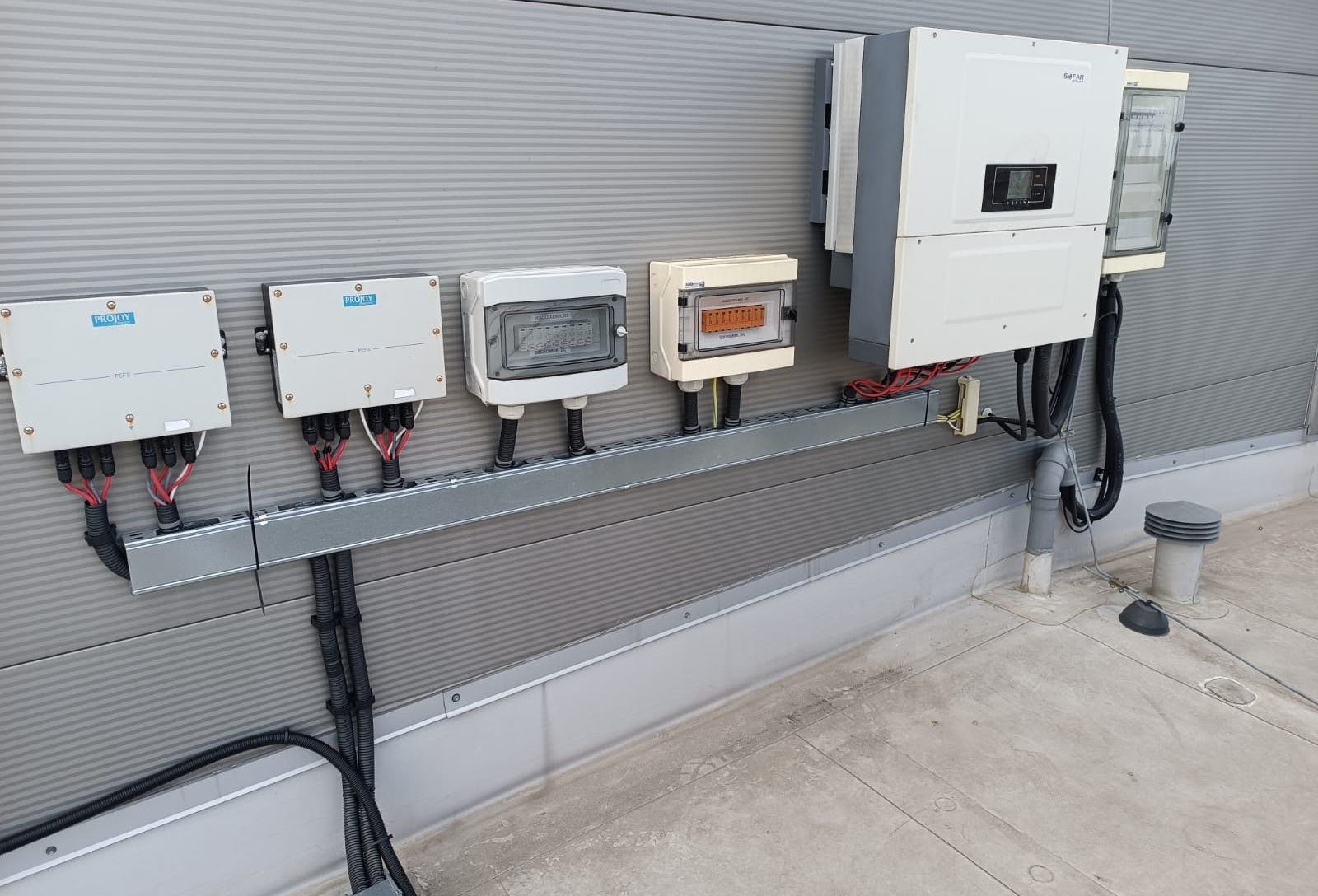 Instalacja elektryczna z trzema skrzynkami przyłączeniowymi Projoy, skrzynką rozdzielczą z przezroczystą pokrywą, skrzynką z bezpiecznikami i falownikiem Safari na szarej ścianie z blachy trapezowej.