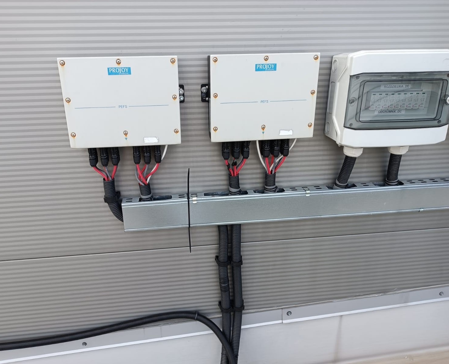 Dwa białe optymalizatory mocy z logo Projoy PEFS oraz rozdzielnica DC zamontowane na szarej ścianie z widocznymi przewodami w peszlach.
