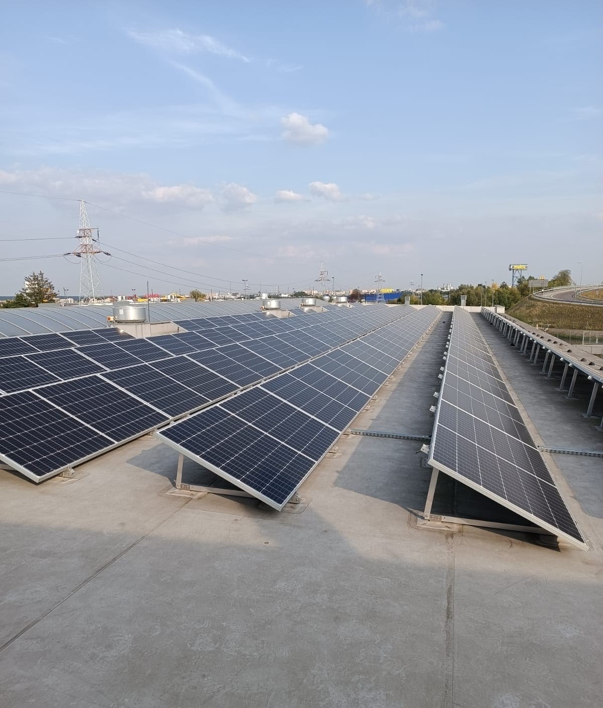 Rozległa instalacja paneli słonecznych na płaskim dachu budynku komercyjnego, widoczne elementy konstrukcyjne i przewody, w tle słupy wysokiego napięcia i autostrada.