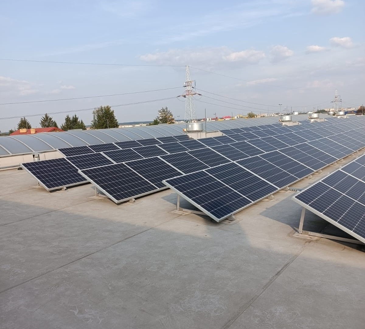 Rząd paneli słonecznych na płaskim dachu budynku przemysłowego, widoczna instalacja wsporcza i słup wysokiego napięcia w tle.